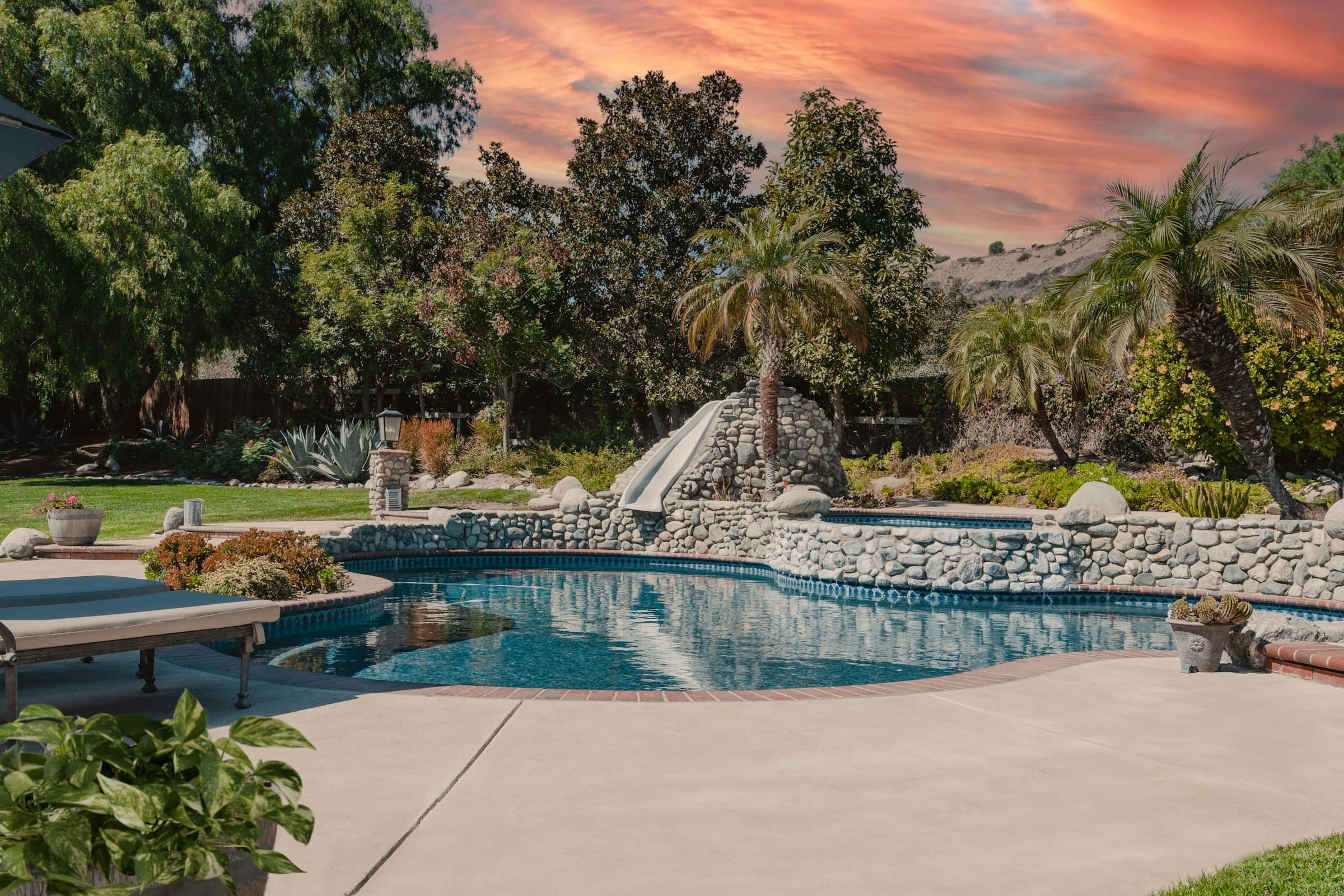 A backyard pool with a rock waterfall and slide at sunset.