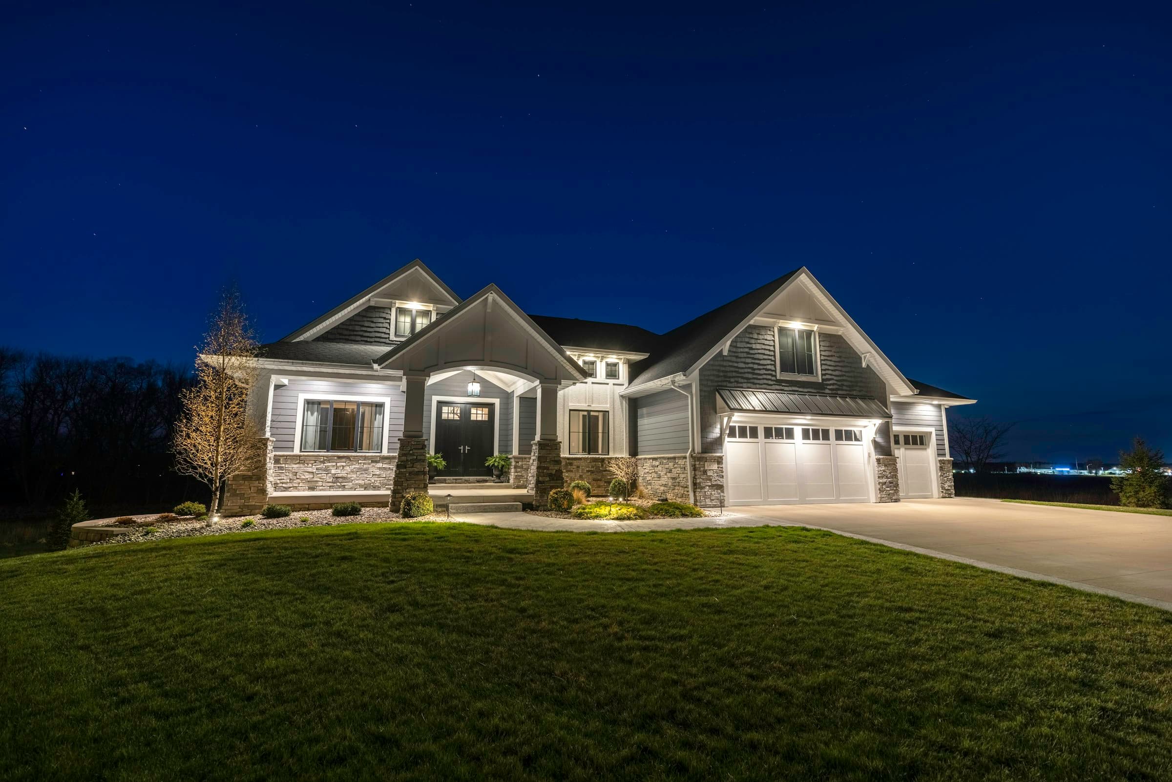 A two-storey craftsman home at blue hour, uplights on the facade and a lit front lawn and drive.