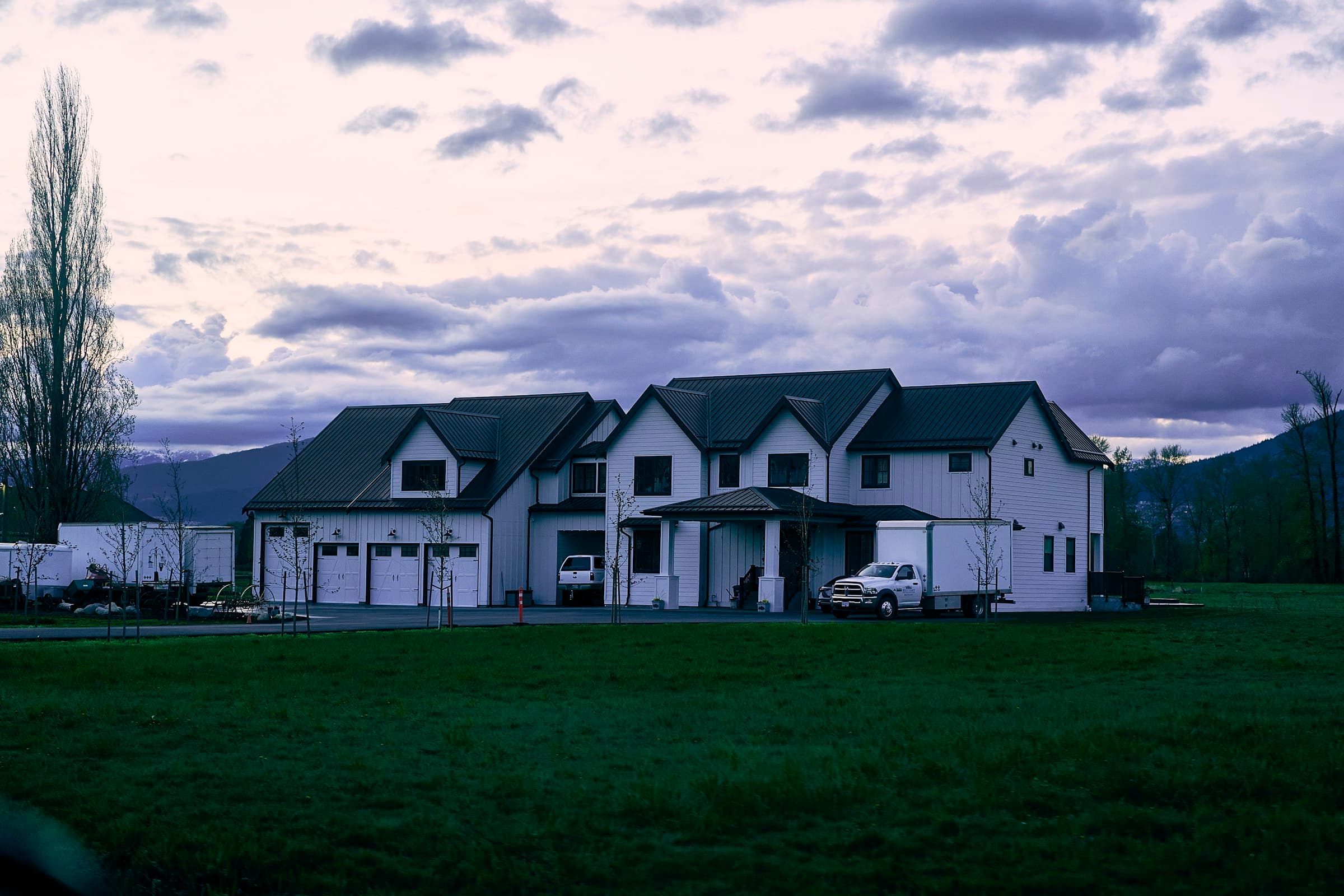 A white modern farmhouse on a wide lawn at dusk with mountains behind.