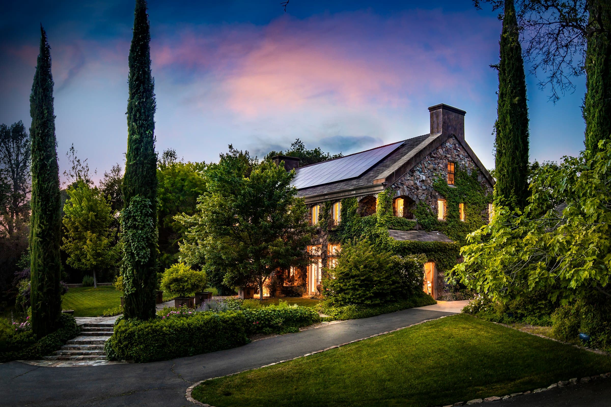 A gray and white home at twilight above a lit, planted front garden.