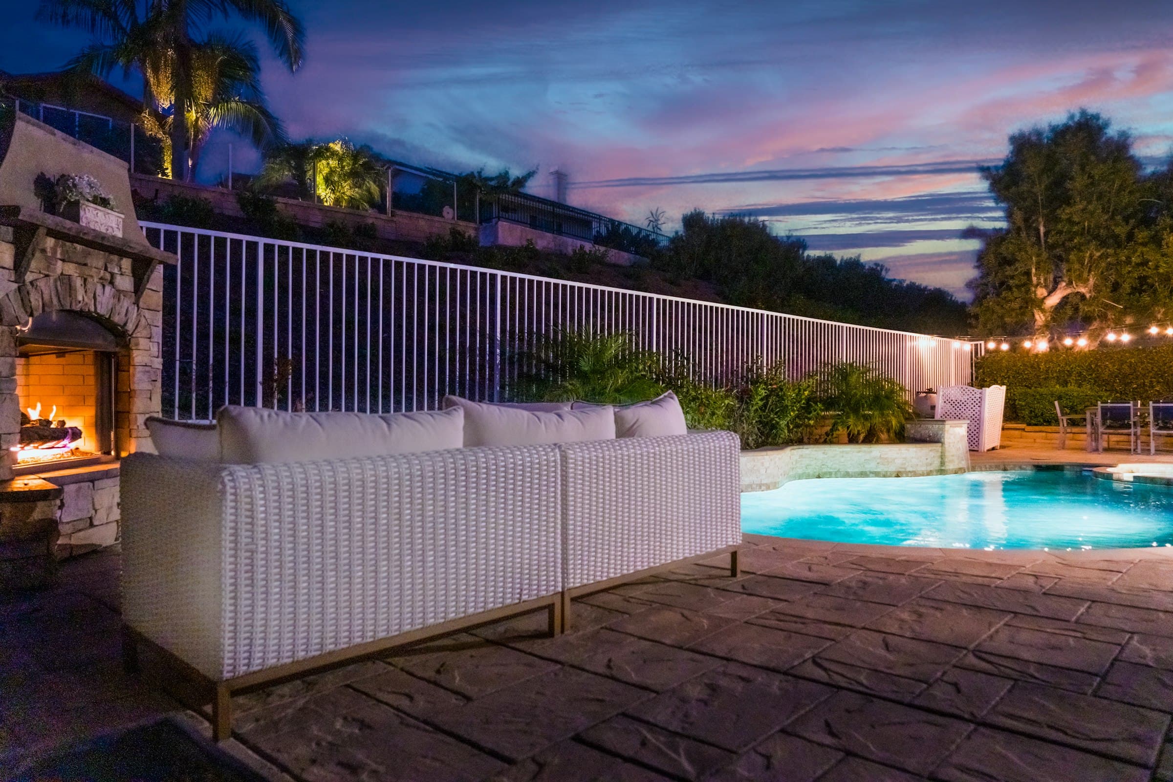 A patio with an outdoor fireplace and seating beside a lit pool at dusk.