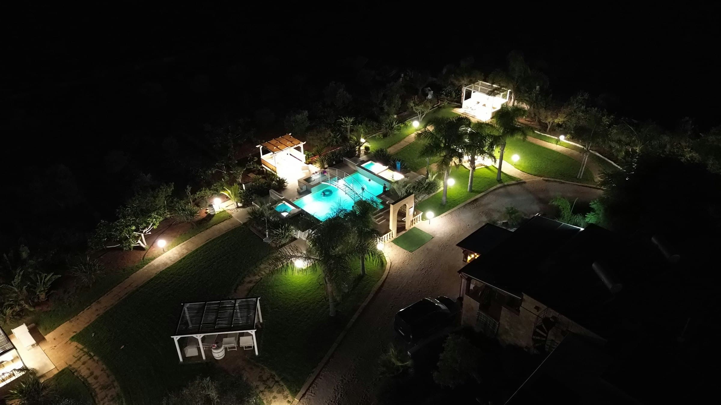 A lit backyard pool and garden at night seen from above, path lights along the lawn.