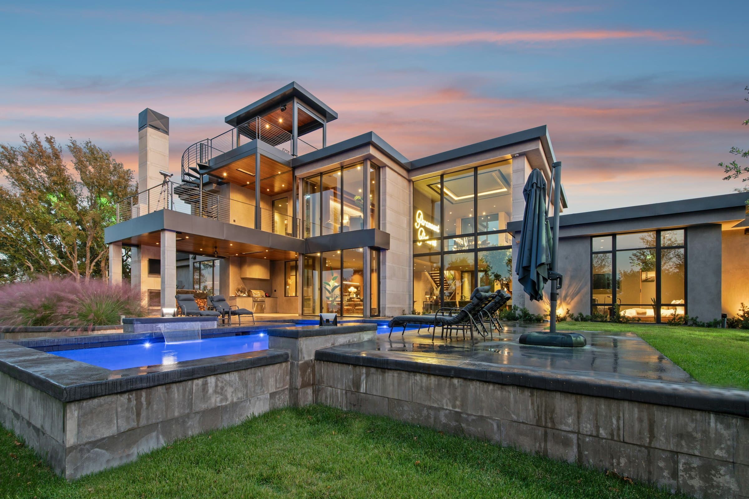 A modern home at dusk, its raised pool and spa lit blue beside the lawn.