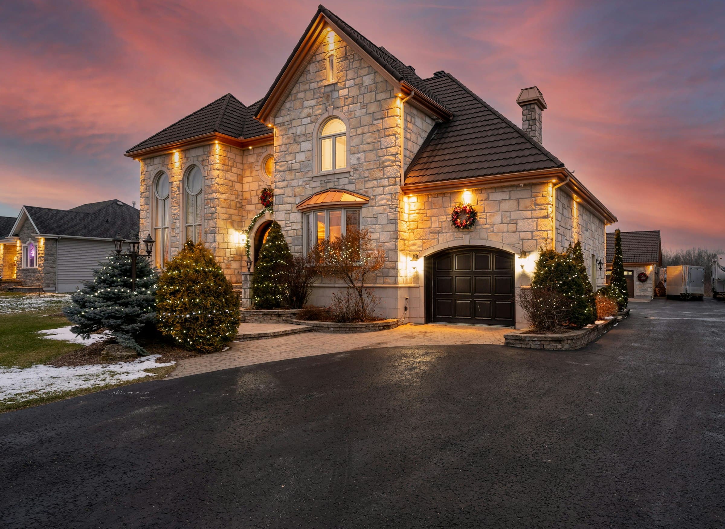 A stone home at dusk seen from the driveway, the facade and garage lit warm.