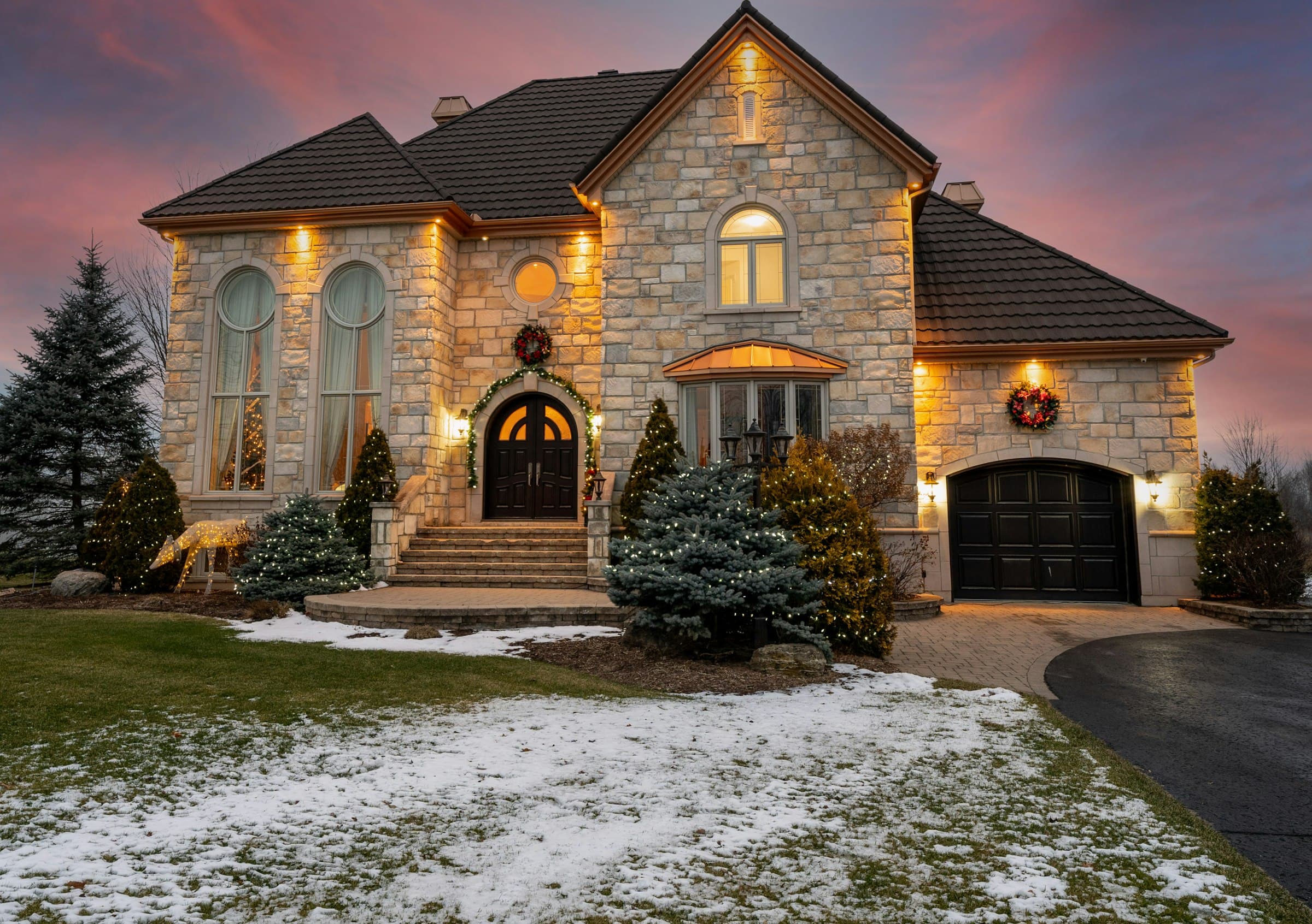 A stone two-storey home at dusk, warm light on the stonework and entry above a front lawn.